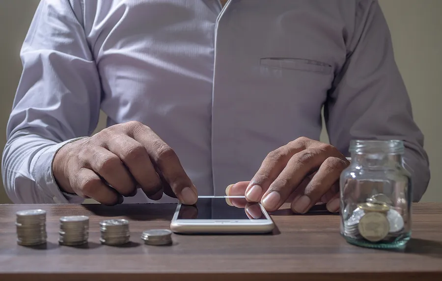 un homme manipulant un téléphone portable posé sur un bureau, à côté d'un bocal contenant des pièces de monnaie et d'une pile de pièces empilées sur la table