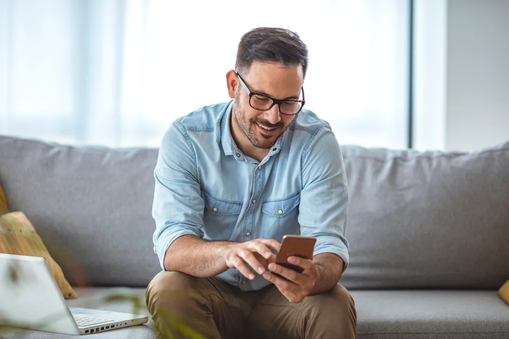 un homme portant des lunettes et une chemise bleue boutonnée, assis sur un canapé gris, souriant à son téléphone portable qu'il tient dans ses mains