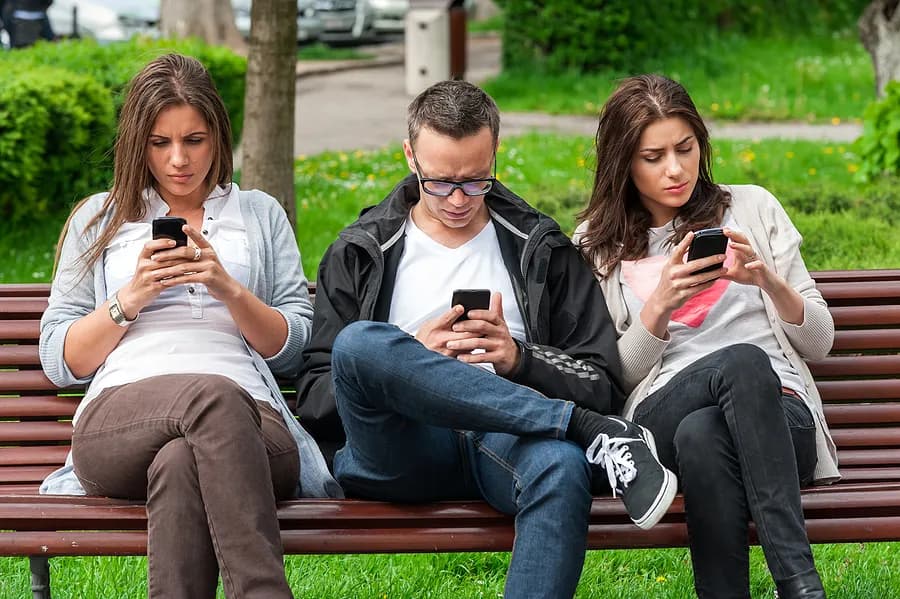 Trois personnes assises sur un banc dans un parc, occupées à consulter leur téléphone portable.