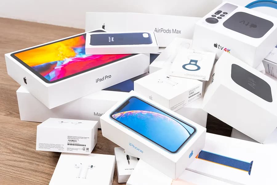 Une pile de boîtes de produits Apple, notamment des iPhone, des iPad Pro, des AirPods, des Apple Watch et des Apple TV, disposées sur une surface en bois.