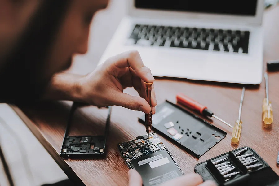Un technicien utilise un tournevis de précision pour réparer les composants internes d'un smartphone sur un bureau en bois, entouré de petits outils, de pièces de rechange et d'un ordinateur portable ouvert.
