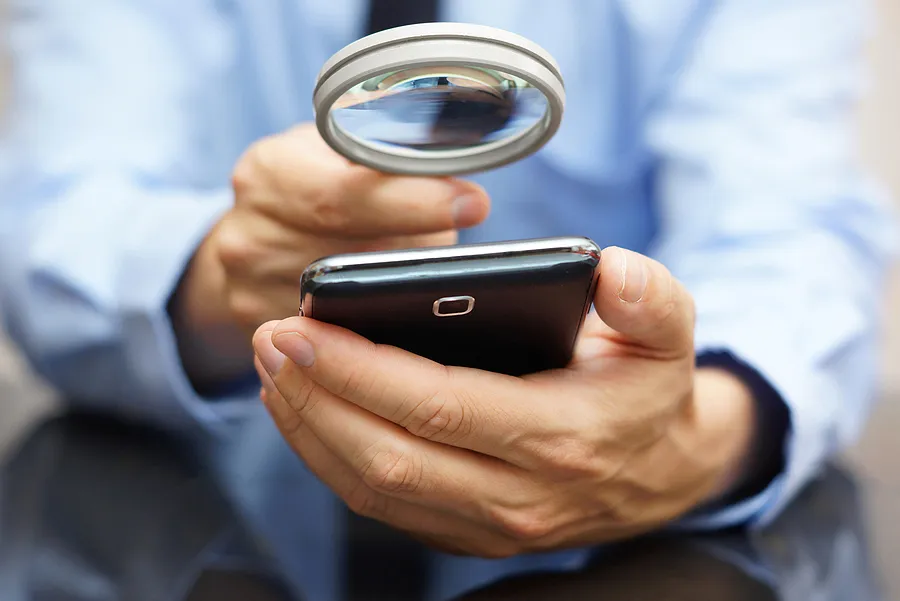 Une personne examinant attentivement un smartphone à l'aide d'une loupe, afin de vérifier son authenticité ou de détecter d'éventuels problèmes.