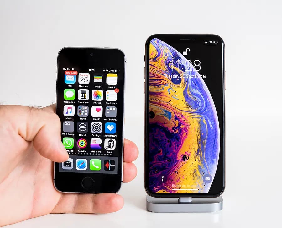 Une main tenant un petit iPhone d'ancienne génération à côté d'un iPhone plus grand et plus récent posé sur un support, illustrant les différences de taille et de design entre un ancien iPhone et un modèle plus récent.