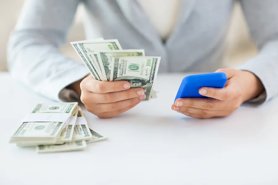 Personne tenant un smartphone dans une main et de l'argent liquide dans l'autre, illustrant la valeur de revente du téléphone et la vente d'un téléphone d'occasion contre de l'argent.